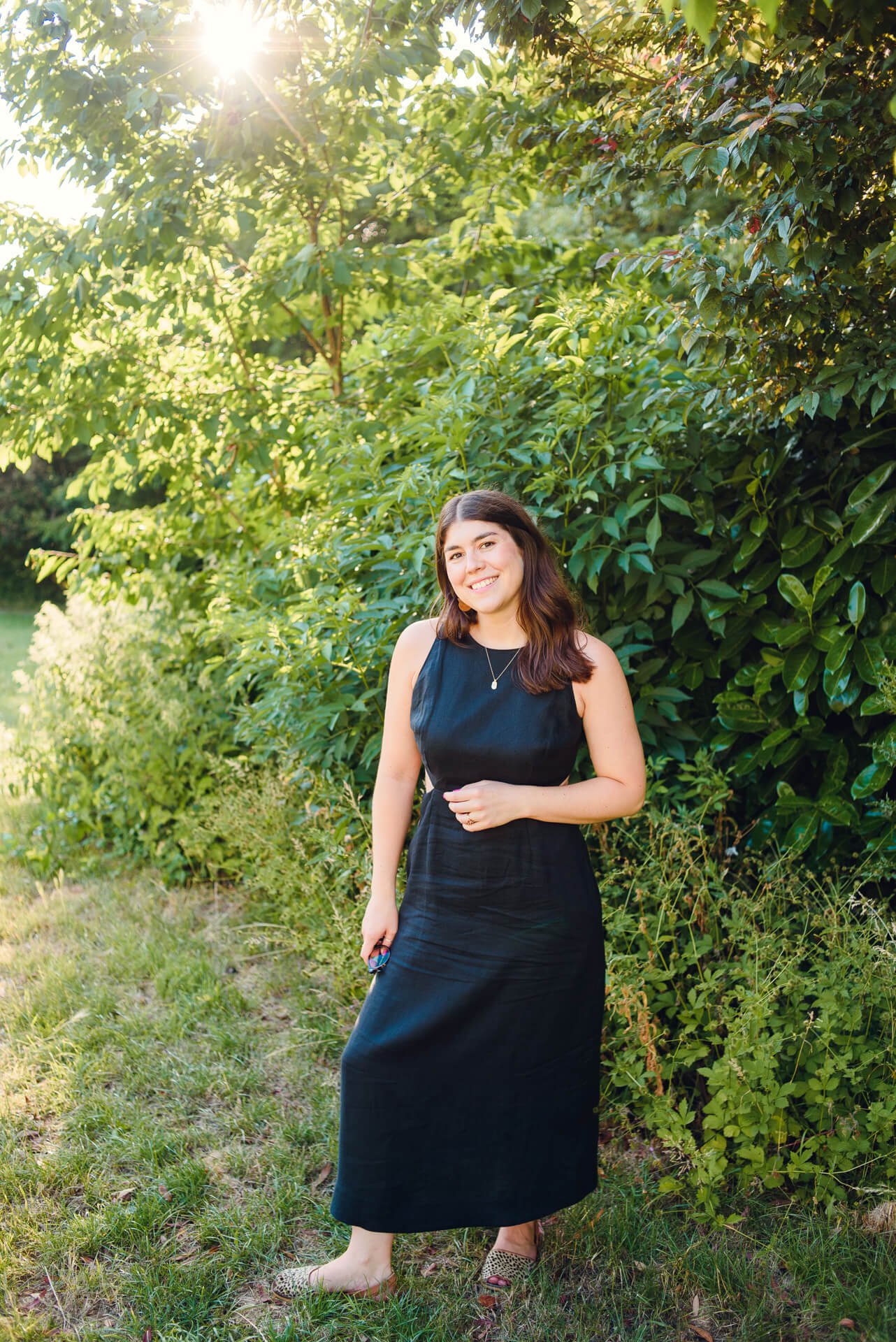 Brown-haired woman standing in a sunny park smiling.